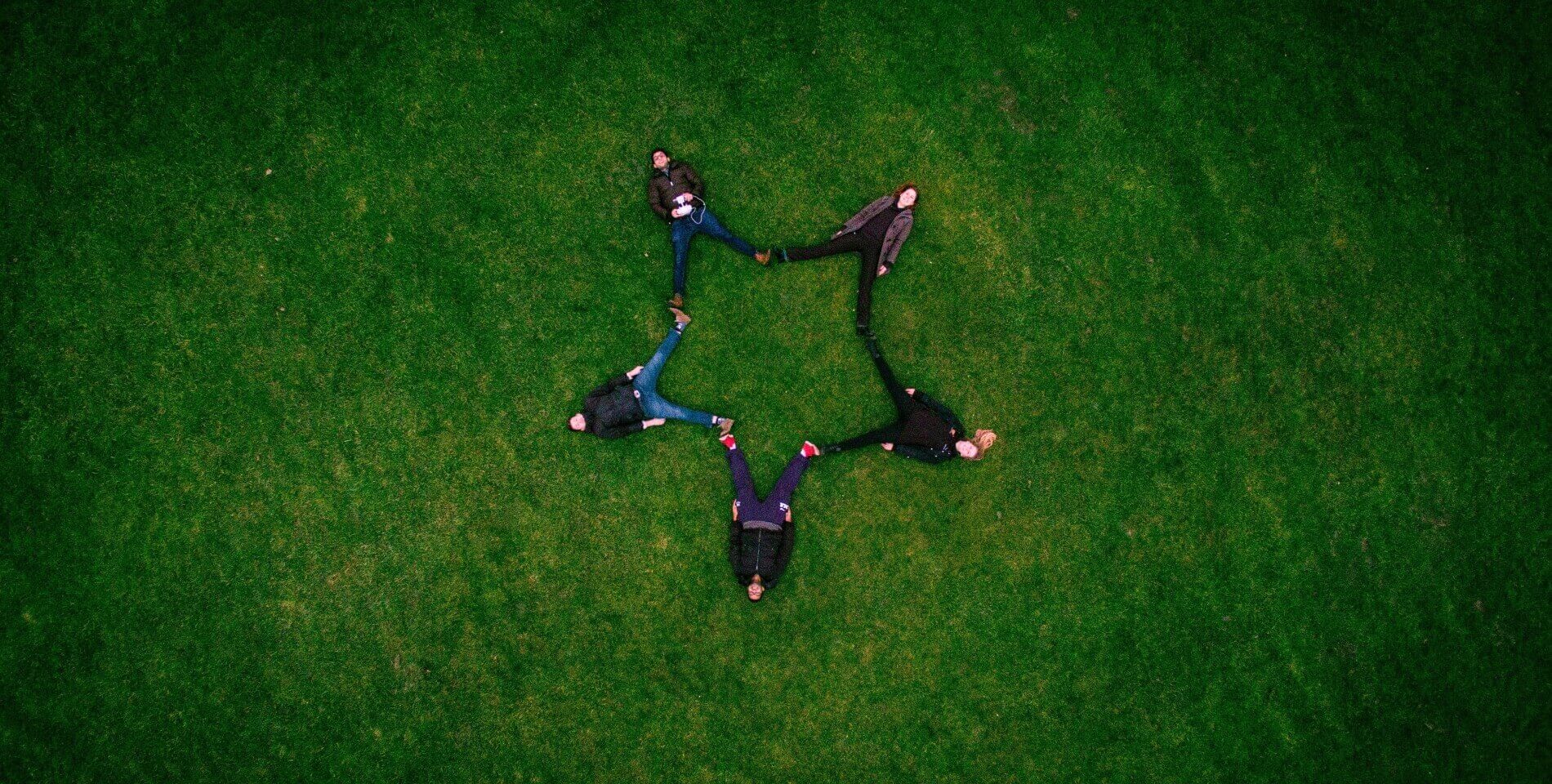 Aerial view of five people laying on green grass area joining their feet to form a star shape